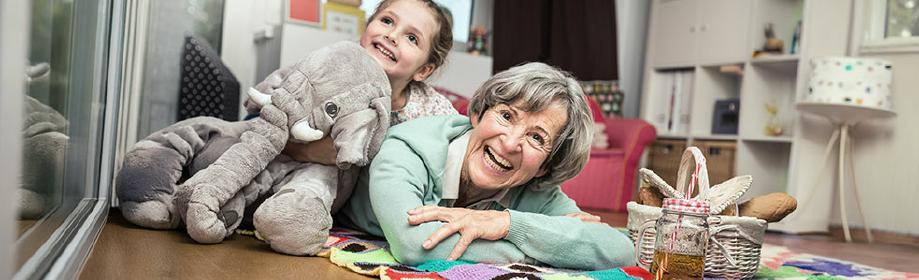 Ältere Frau und Kind liegen lachend auf einem Teppich, Kuscheltier und Picknickkorb im Vordergrund